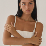 Against a plain background, a woman with long black hair and a white top stands with arms crossed, showcasing gold jewelry from Twenty Compass, including the Antibes Earrings - Gold.