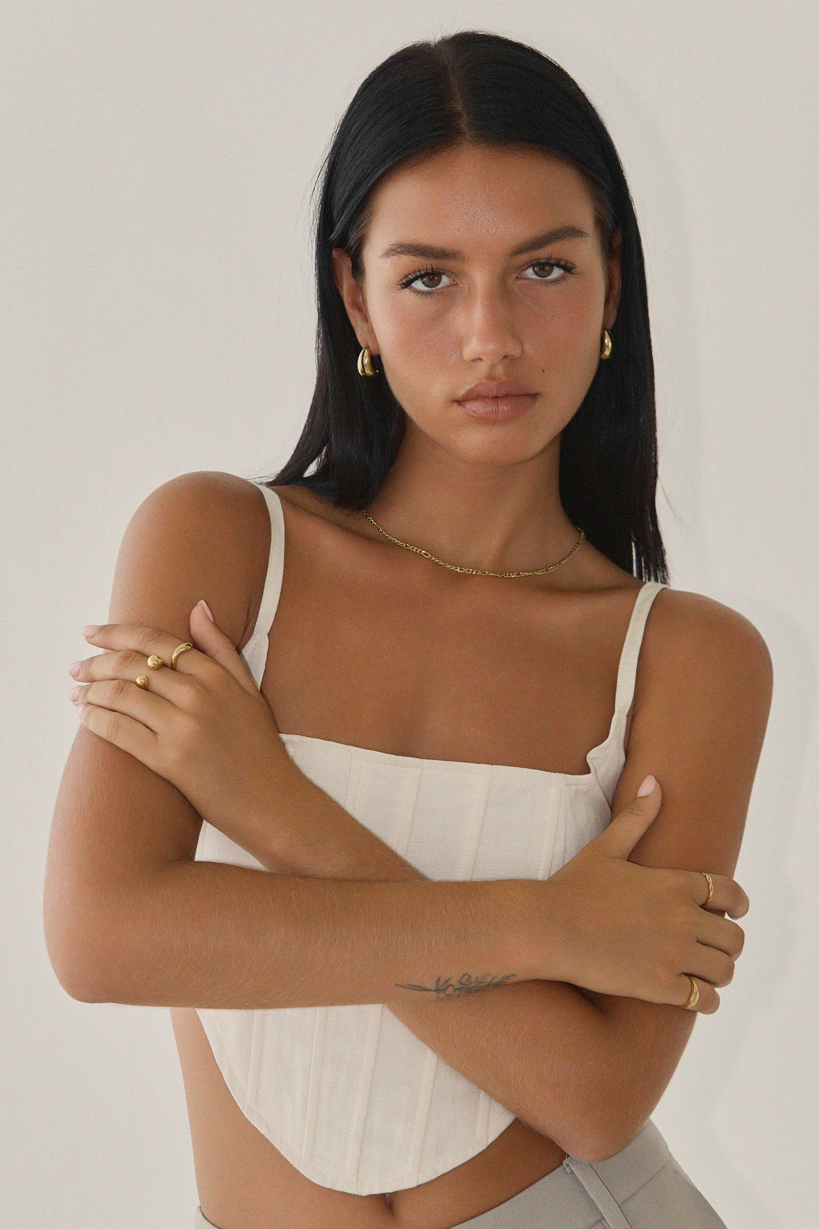 Against a plain background, a woman with long black hair and a white top stands with arms crossed, showcasing gold jewelry from Twenty Compass, including the Antibes Earrings - Gold.