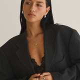 A person with long black hair wears a large black blazer over a lace top, accessorized with timeless studs and Twenty Compass Antibes Earrings - Silver. They stand against a plain background, gazing slightly to the side, embodying the essence of a capsule wardrobe.