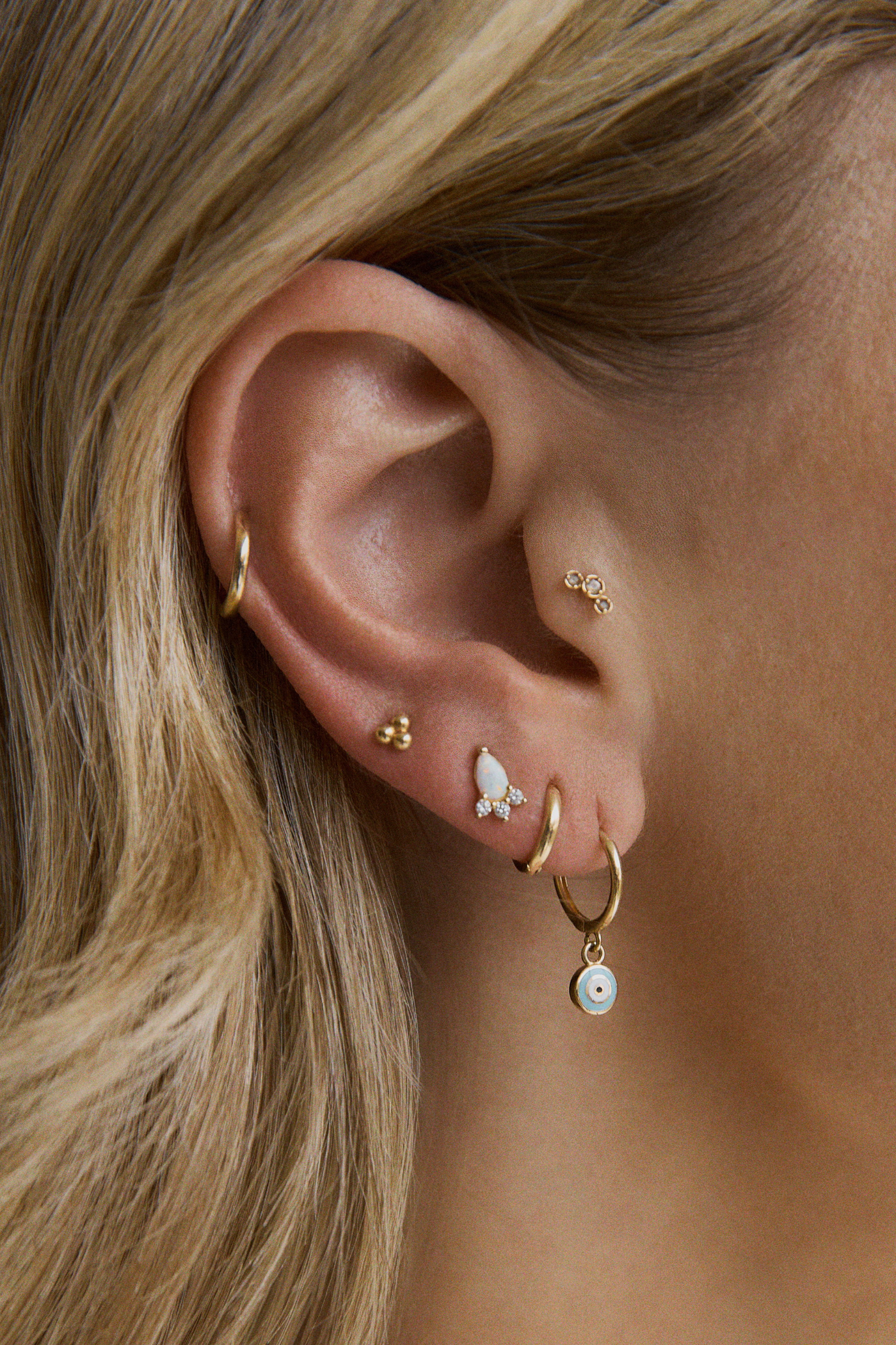 Close-up of a woman's ear with multiple gold earrings: a hoop, a teardrop opal stud with cubic zirconia accents, a small cluster of gold studs, and a gold hoop with a dangling eye charm Boucle d'Oreilles Opale - Or 10 Carats