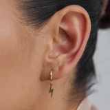 Close-up of a woman's ear wearing a single gold huggie hoop earring with a dangling gold lightning bolt charm.