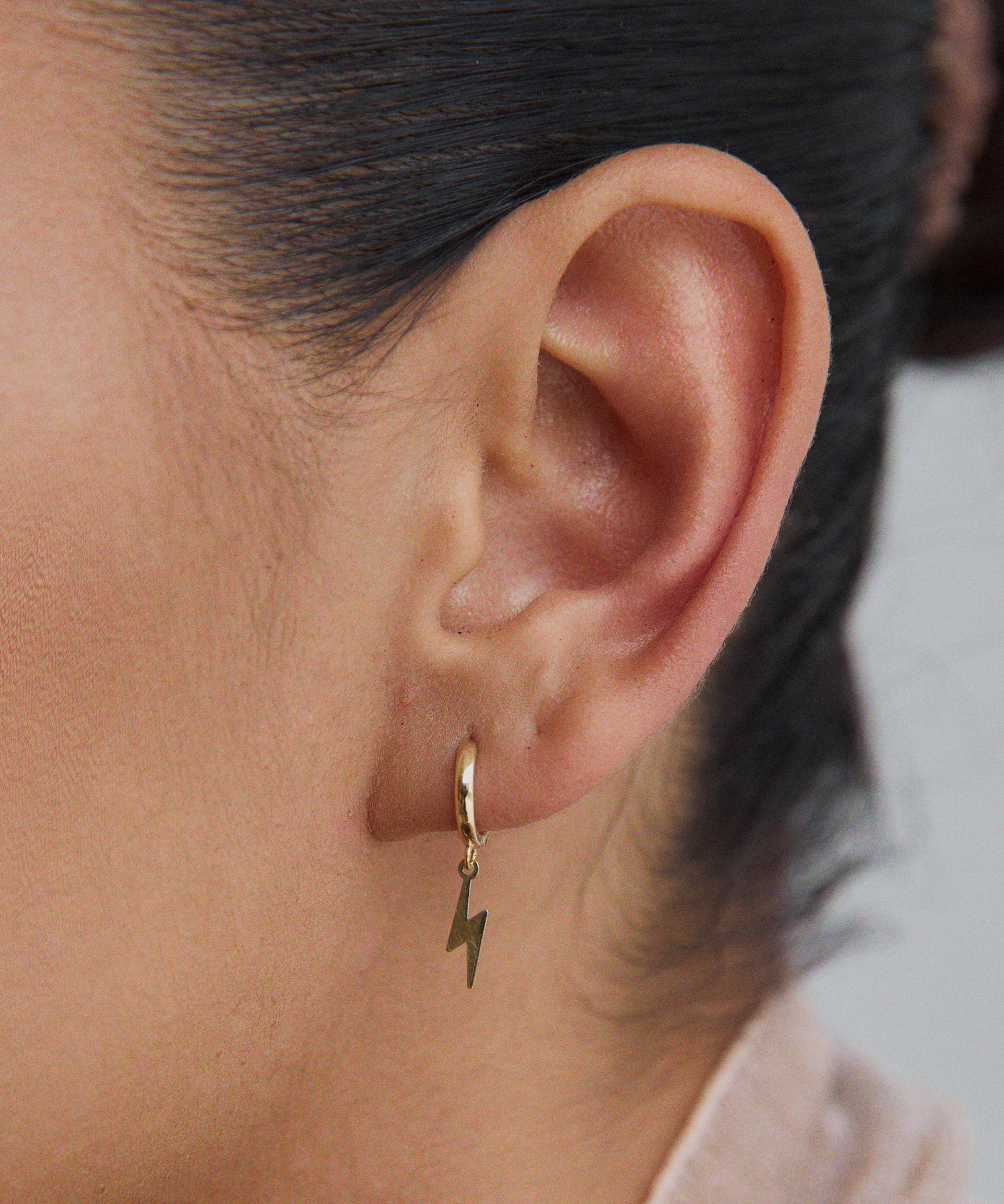 Close-up of a woman's ear wearing a single gold huggie hoop earring with a dangling gold lightning bolt charm.