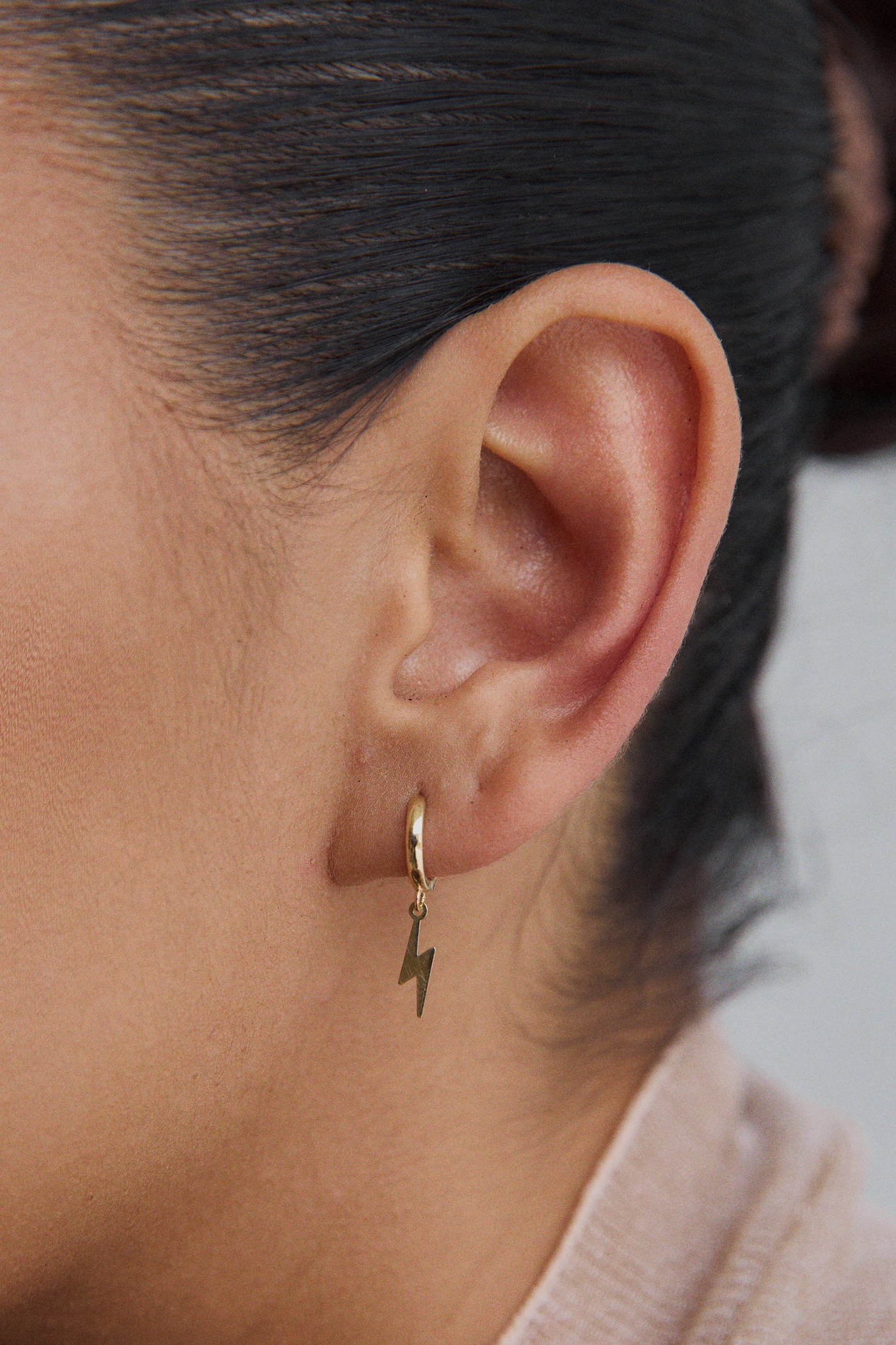 Close-up of a woman's ear wearing a single gold huggie hoop earring with a dangling gold lightning bolt charm. Boucles d'Oreilles Éclair - Or 10 Carats