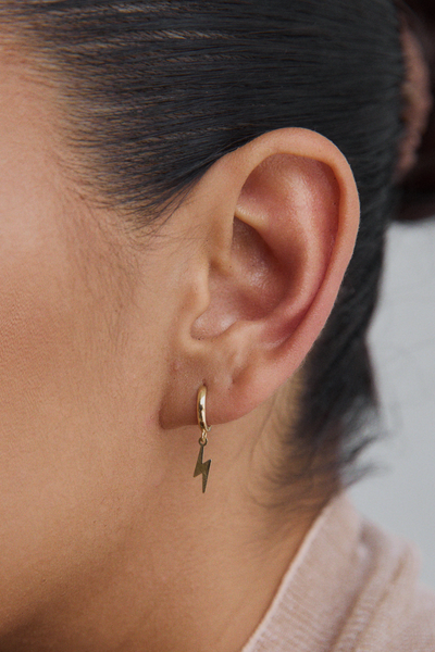 Close-up of a woman's ear wearing a single gold huggie hoop earring with a dangling gold lightning bolt charm.