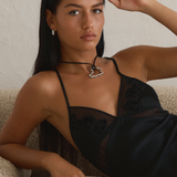 A woman with long dark hair lounges on a beige couch, wearing a black dress with sheer accents and silver Antibes Earrings by Twenty Compass. Her neutral expression is visible as she rests her head on her hand, gazing towards the camera.
