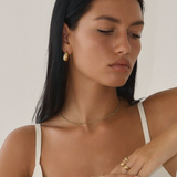 A woman with long dark hair in a white sleeveless top, Antibes Earrings - Gold by Twenty Compass, a delicate gold necklace, and multiple gold rings poses with her eyes closed. She exudes calm sophistication against a plain backdrop, embodying the essence of a capsule wardrobe.