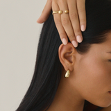 A person with long black hair, wearing Twenty Compass Antibes Earrings - Gold, a gold necklace, and two gold rings, holds their head with one hand. Their nails are painted light pink. The plain background highlights the elegance of these capsule wardrobe accessories.
