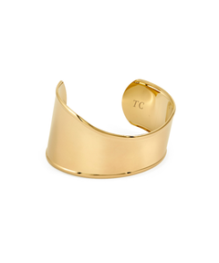 A wide, polished gold statement cuff bracelet featuring a smooth, asymmetrical wavy design and an open back, with the letters "TC" subtly engraved on the inner curve, isolated on a white background.