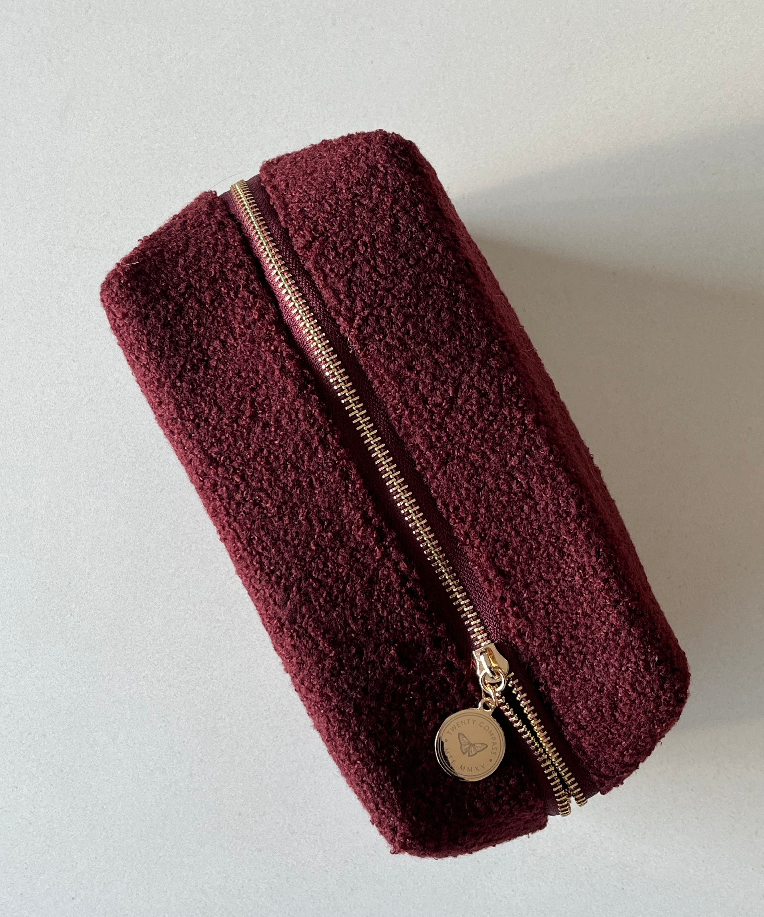 A dark red travel pouch with a gold zipper and a gold pendant with an engraved butterfly design and Twenty Compass logo.