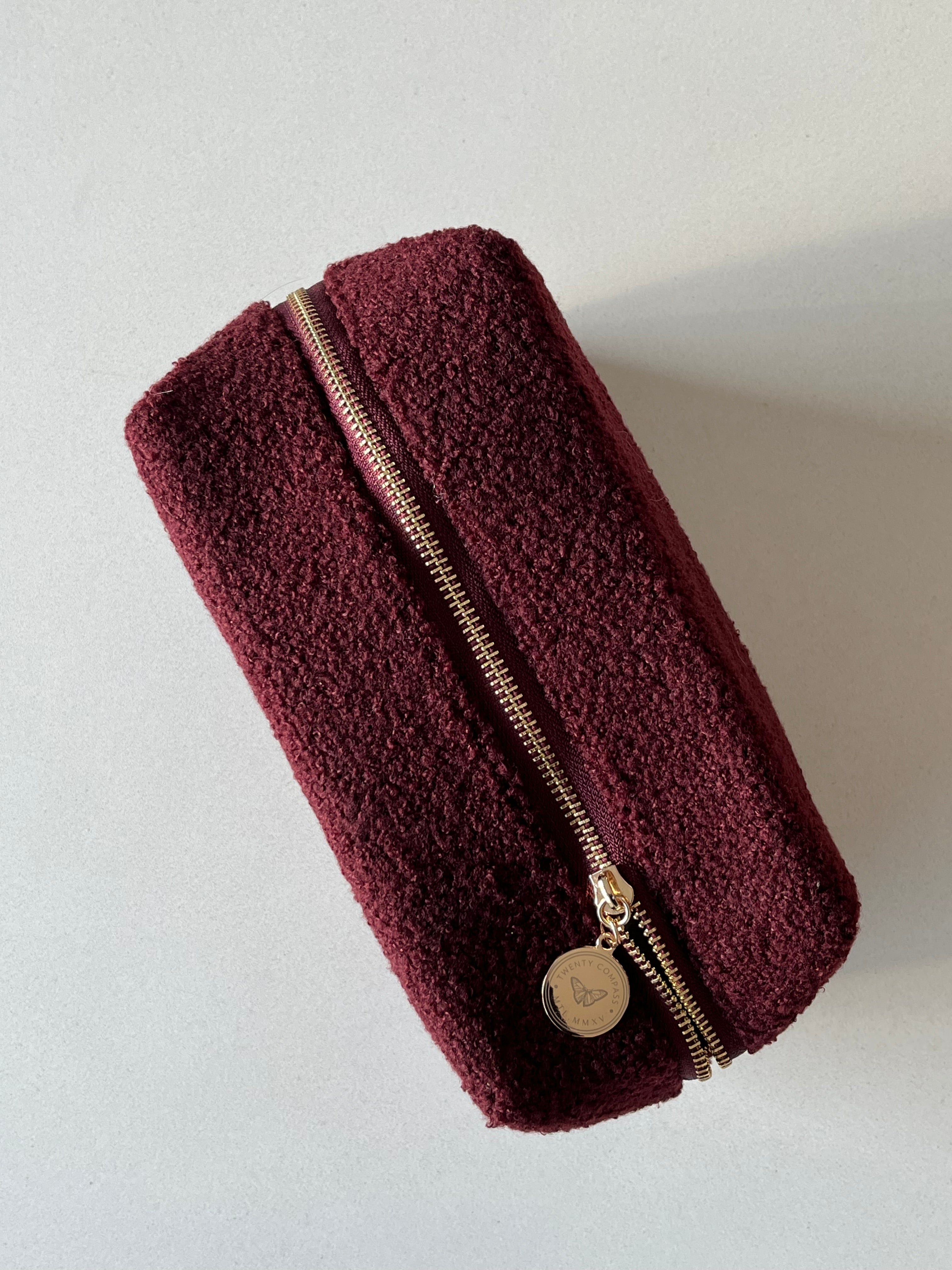 A dark red travel pouch with a gold zipper and a gold pendant with an engraved butterfly design and Twenty Compass logo.