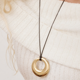 Close-up of a chunky gold donut pendant necklace with a black cord styled on a white ribbed sweater.