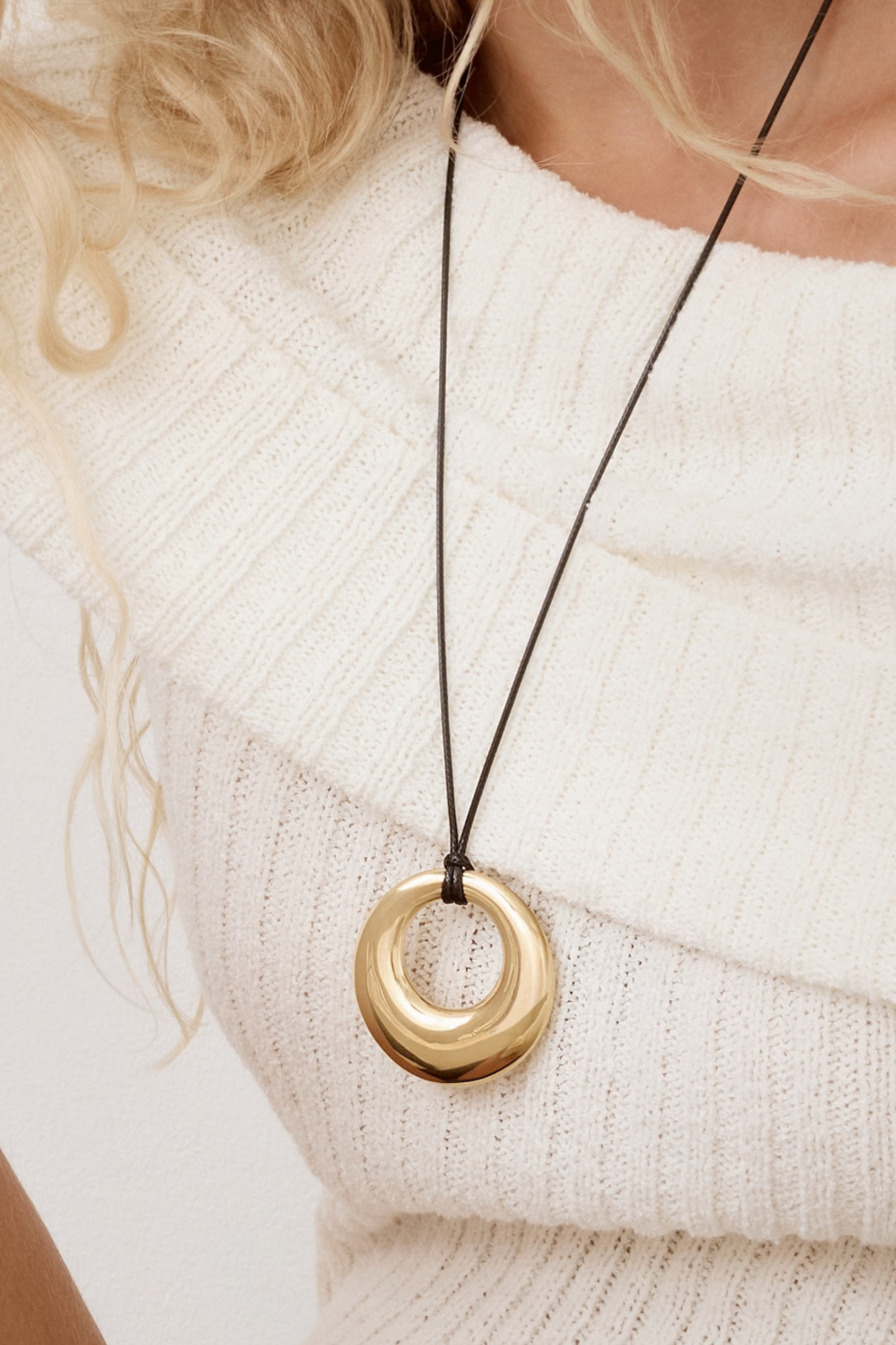 Close-up of a chunky gold donut pendant necklace with a black cord styled on a white ribbed sweater.