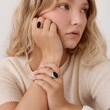 A close-up lifestyle shot of a woman with blonde hair wearing a cream knit sweater and two black rings with oval stone accents.