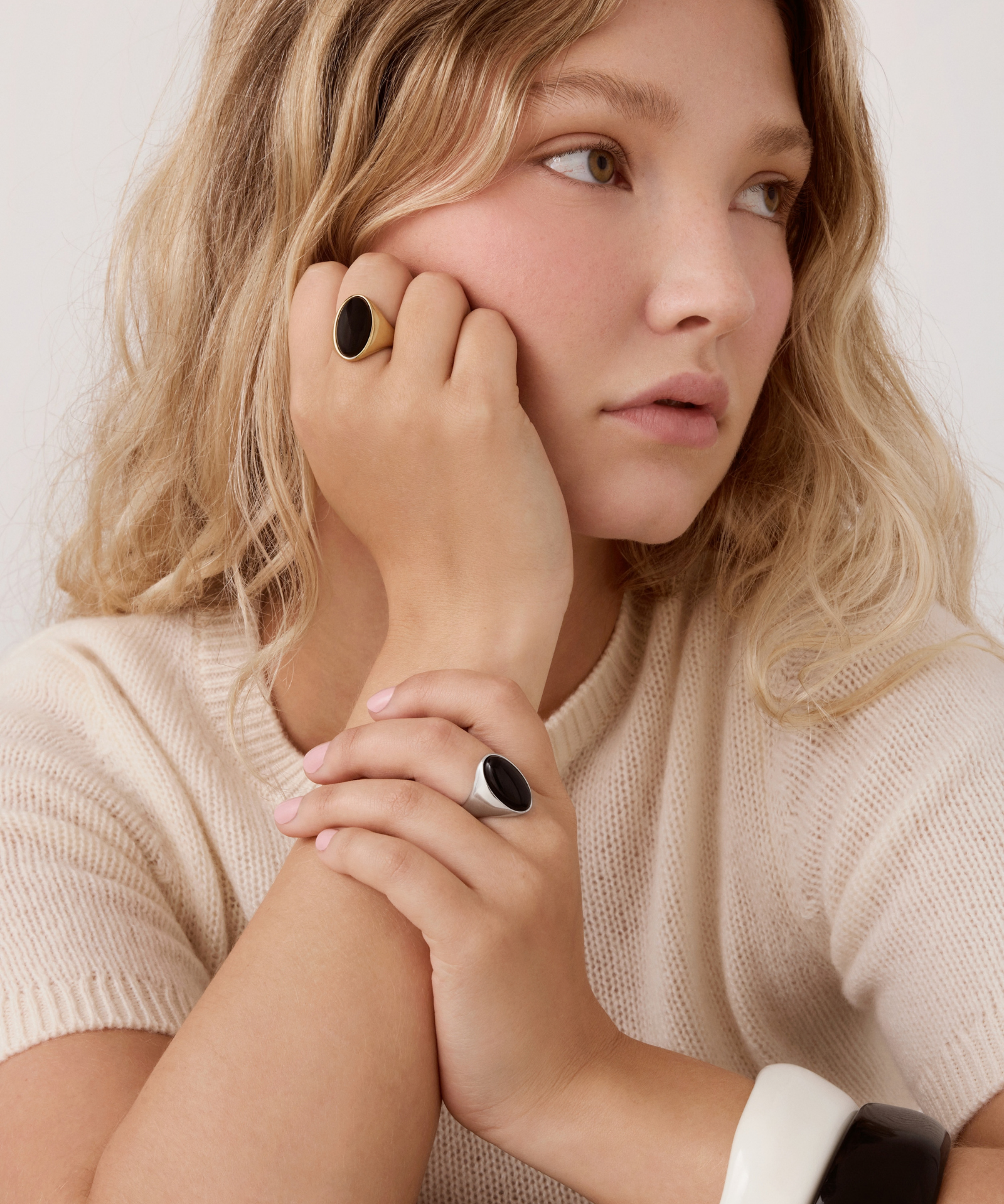 A close-up lifestyle shot of a woman with blonde hair wearing a cream knit sweater and two black rings with oval stone accents.