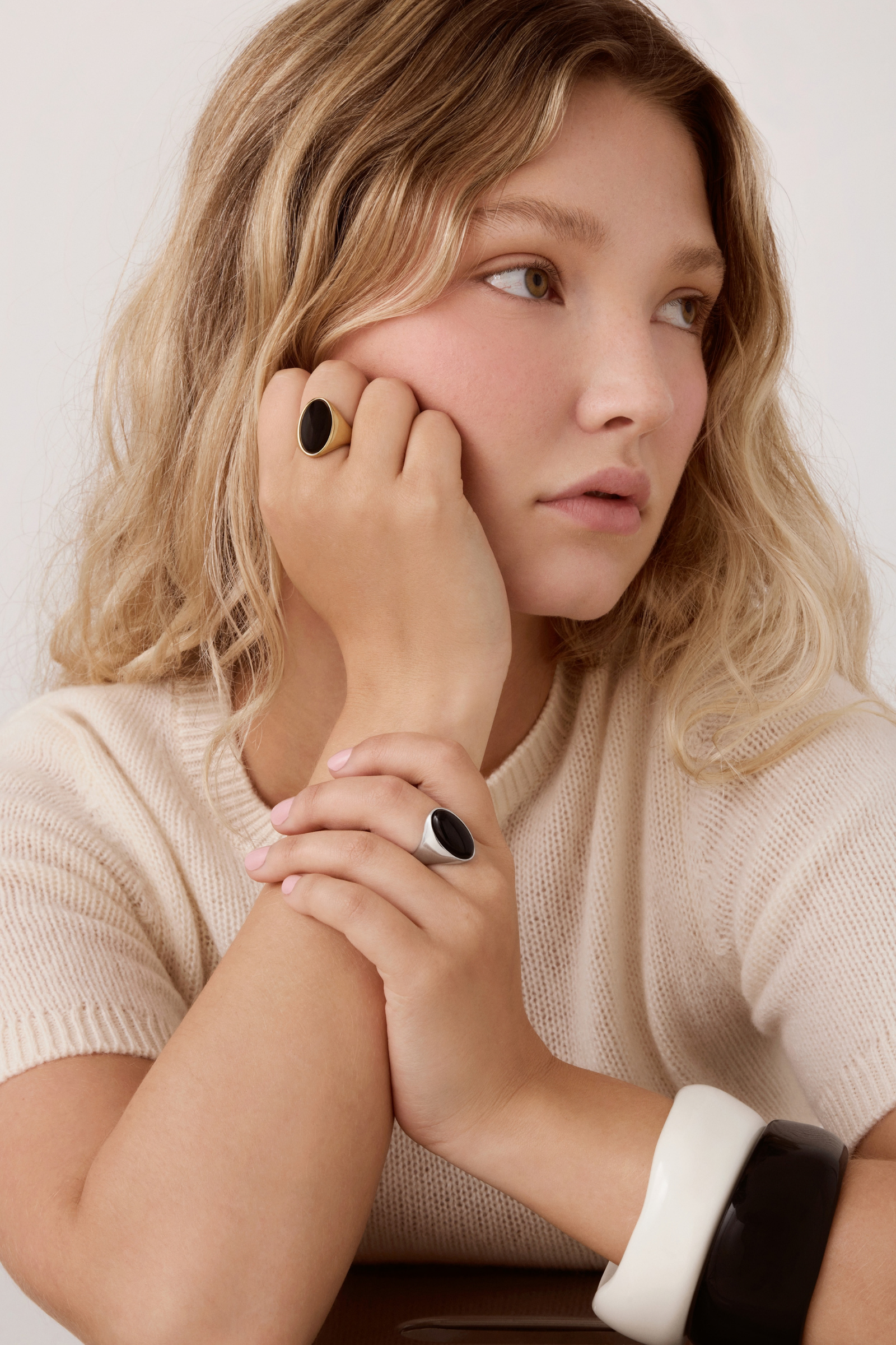 A close-up lifestyle shot of a woman with blonde hair wearing a cream knit sweater and two black rings with oval stone accents.