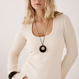 A woman wearing a cream-colored, long-sleeve ribbed scoop-neck top and a black pendant necklace, posing against a plain off-white background.
