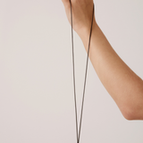 A hand wearing a black ring holds up a long black cord necklace with a large black circular pendant against a plain light beige background.