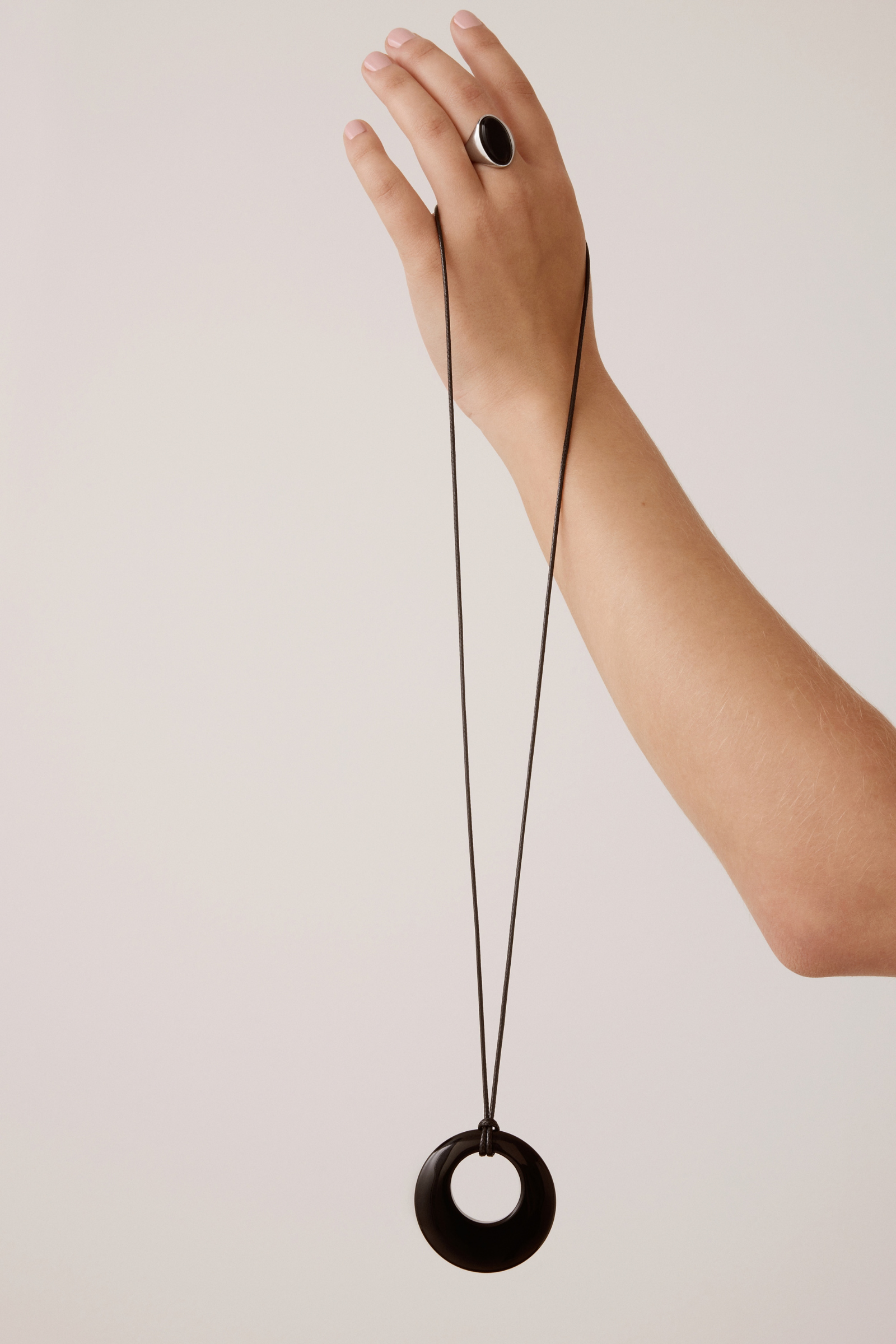 A hand wearing a black ring holds up a long black cord necklace with a large black circular pendant against a plain light beige background.