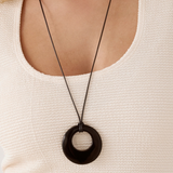 A close-up of a person wearing a long black cord necklace with a large black circular pendant over a cream-colored textured top.