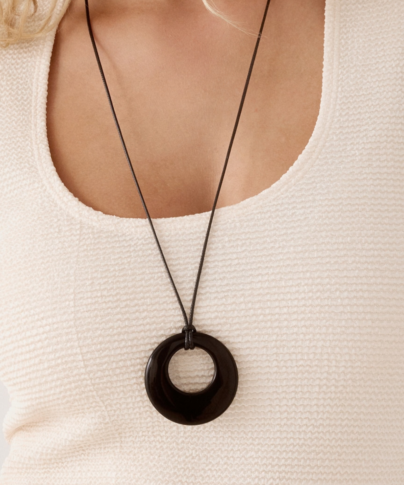 A close-up of a person wearing a long black cord necklace with a large black circular pendant over a cream-colored textured top.