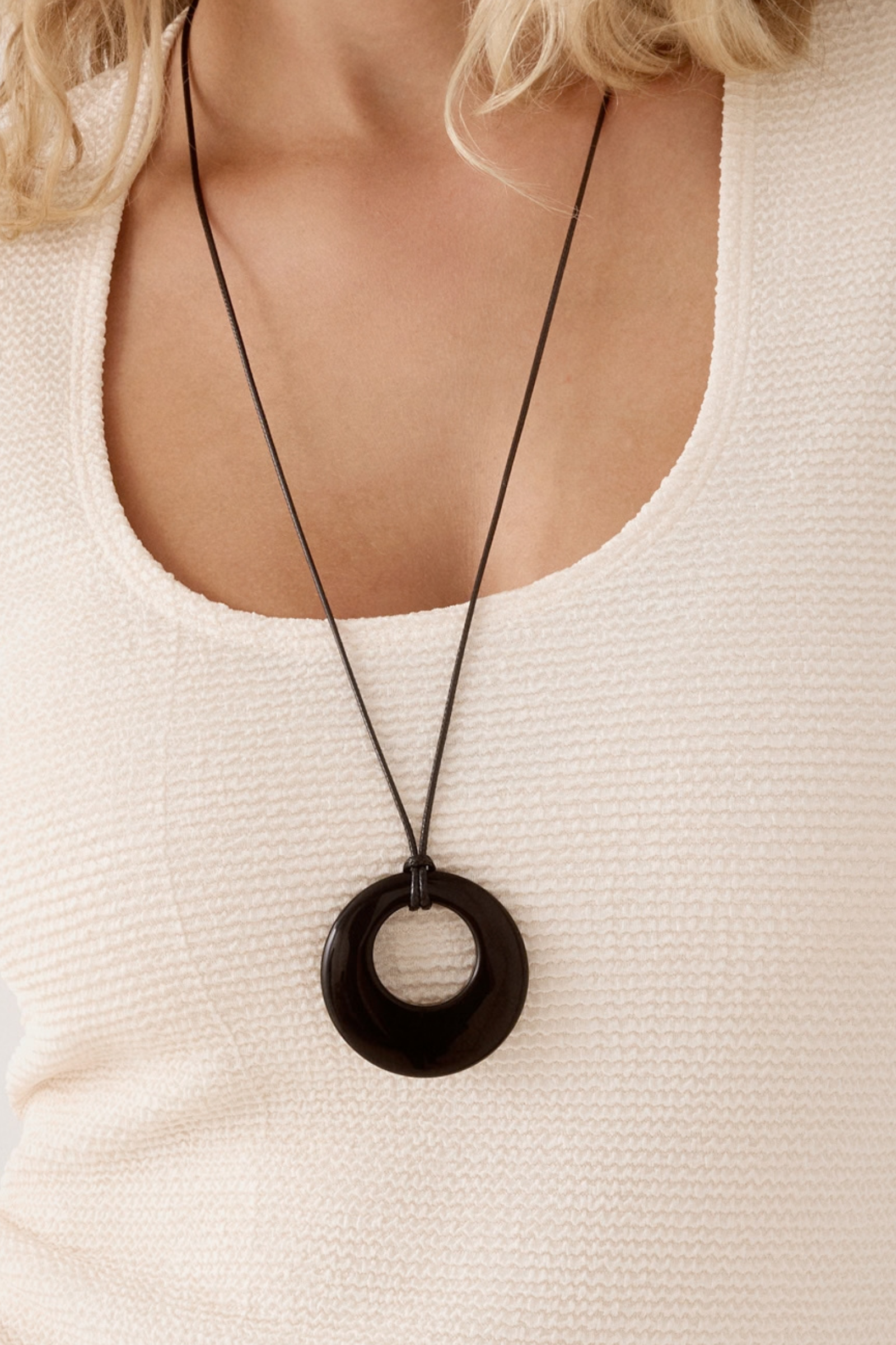 A close-up of a person wearing a long black cord necklace with a large black circular pendant over a cream-colored textured top.