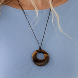 A close-up of a tiger eye gemstone open-circle pendant on a thin dark cord over a light blue top.