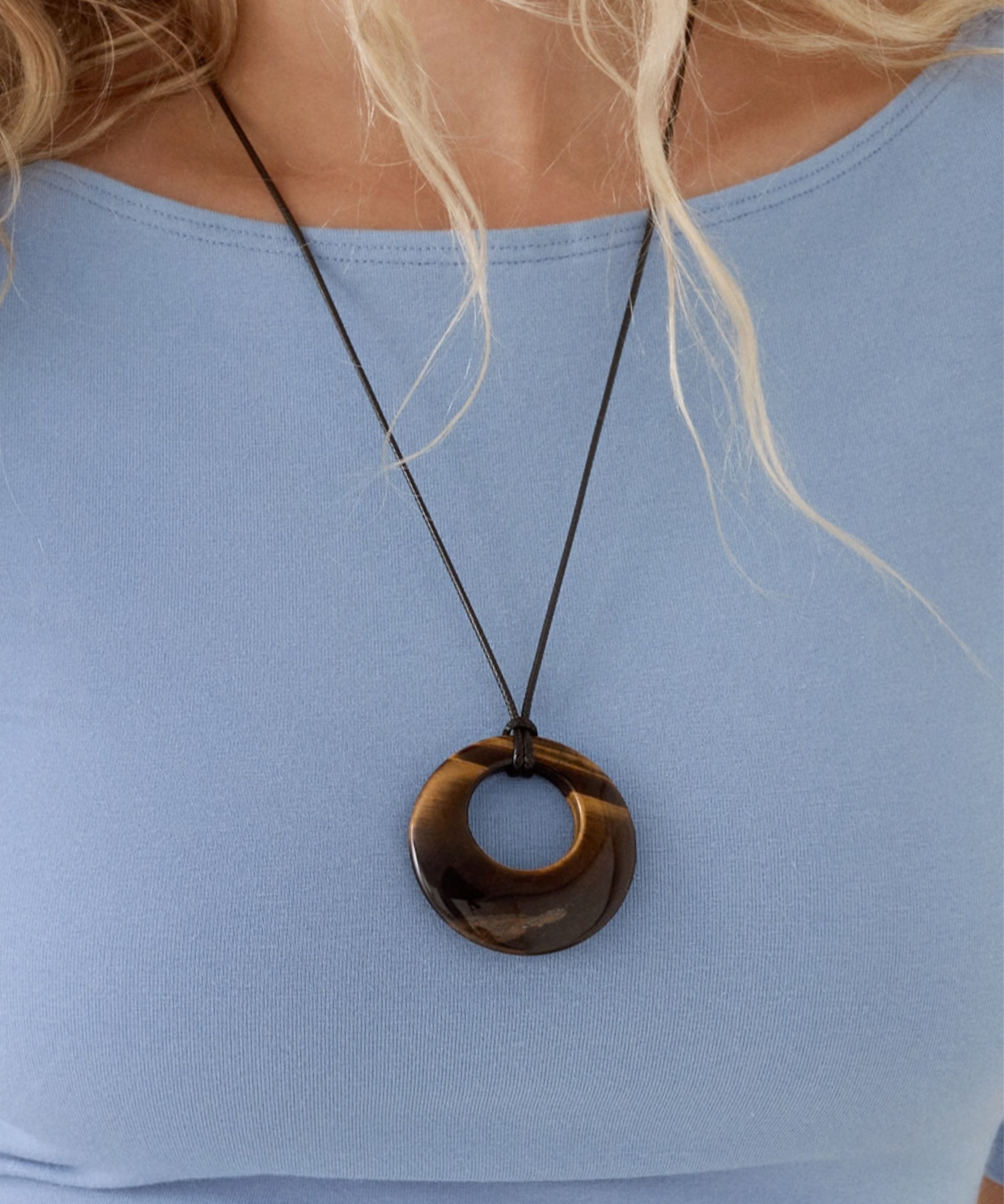 A close-up of a tiger eye gemstone open-circle pendant on a thin dark cord over a light blue top.