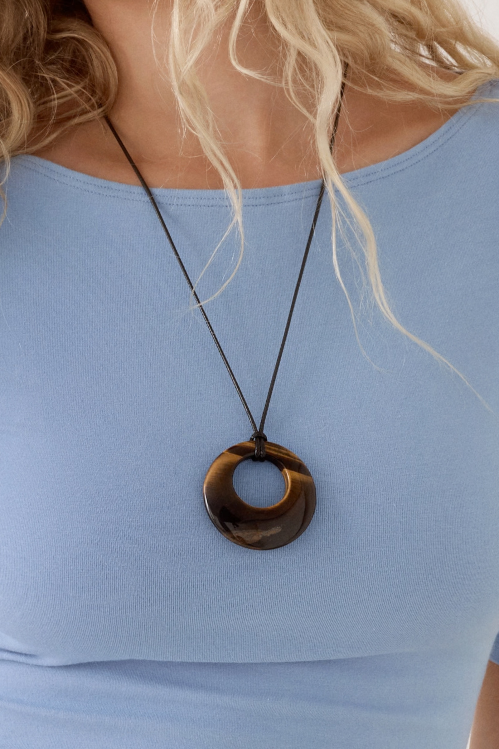 A close-up of a tiger eye gemstone open-circle pendant on a thin dark cord over a light blue top.