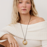 A woman wearing a chunky gold ring pendant necklace on a black cord, styled with an off-the-shoulder cream knit sweater against a light gray background.