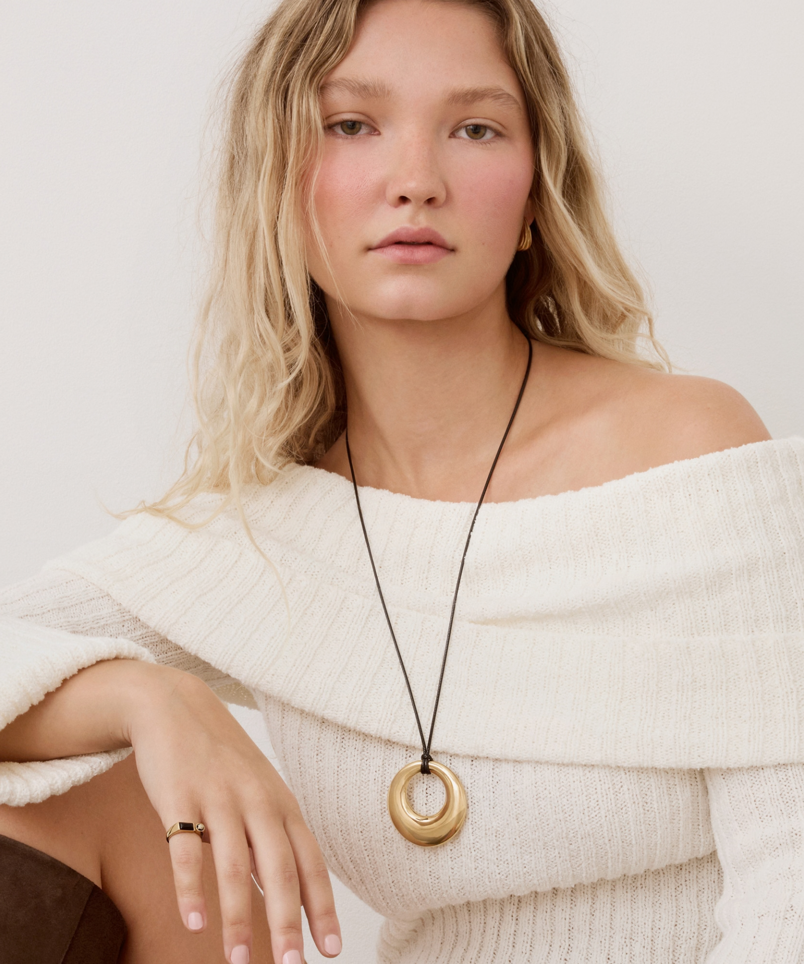 A woman wearing a chunky gold ring pendant necklace on a black cord, styled with an off-the-shoulder cream knit sweater against a light gray background.