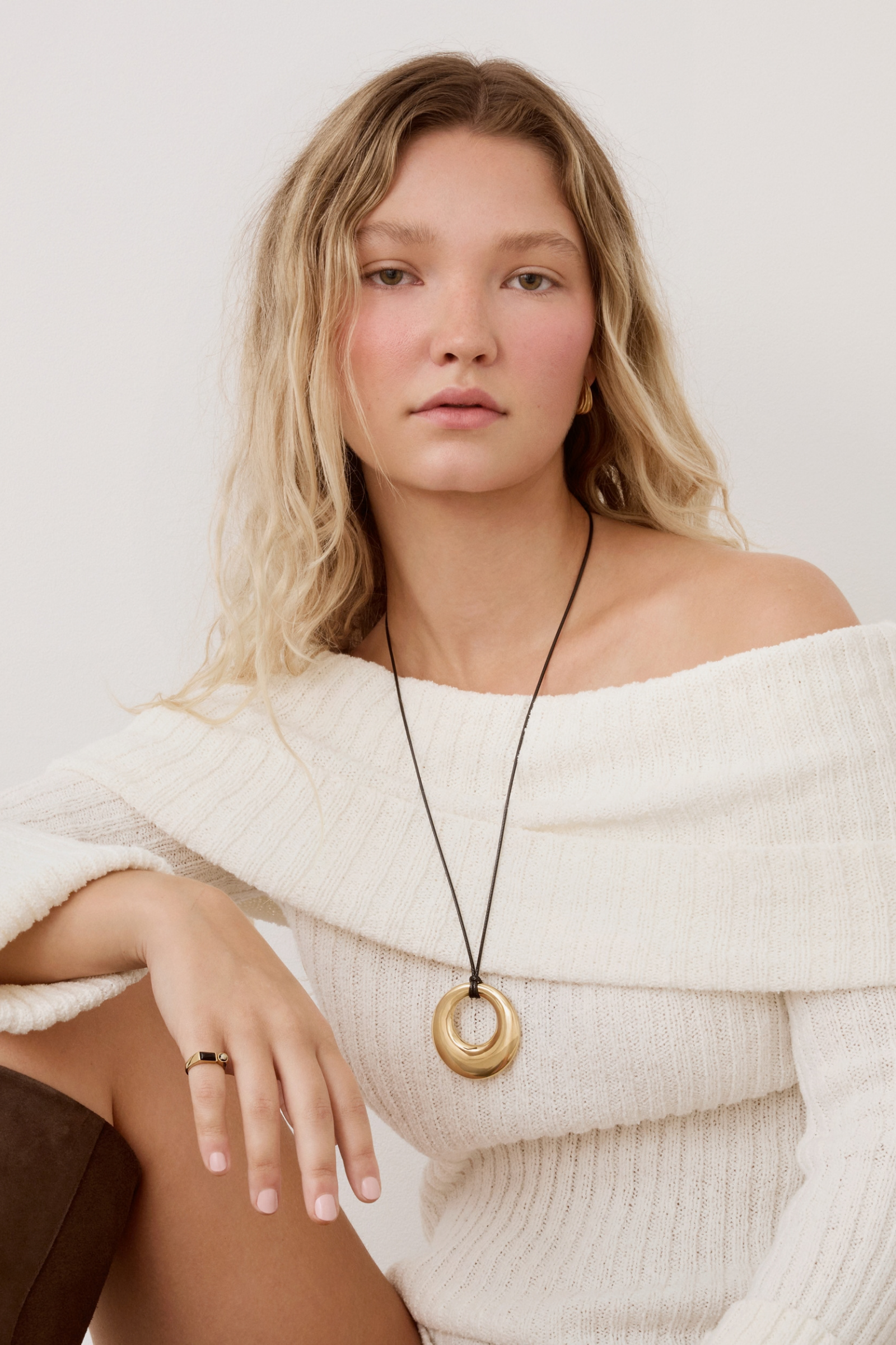 A woman wearing a chunky gold ring pendant necklace on a black cord, styled with an off-the-shoulder cream knit sweater against a light gray background.