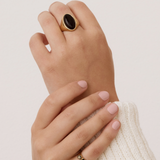 A close-up of a person's hands wearing gold rings, with the larger ring featuring a dark oval stone on the back hand and a smaller gold band with a black stone on the front hand, set against a plain light background.