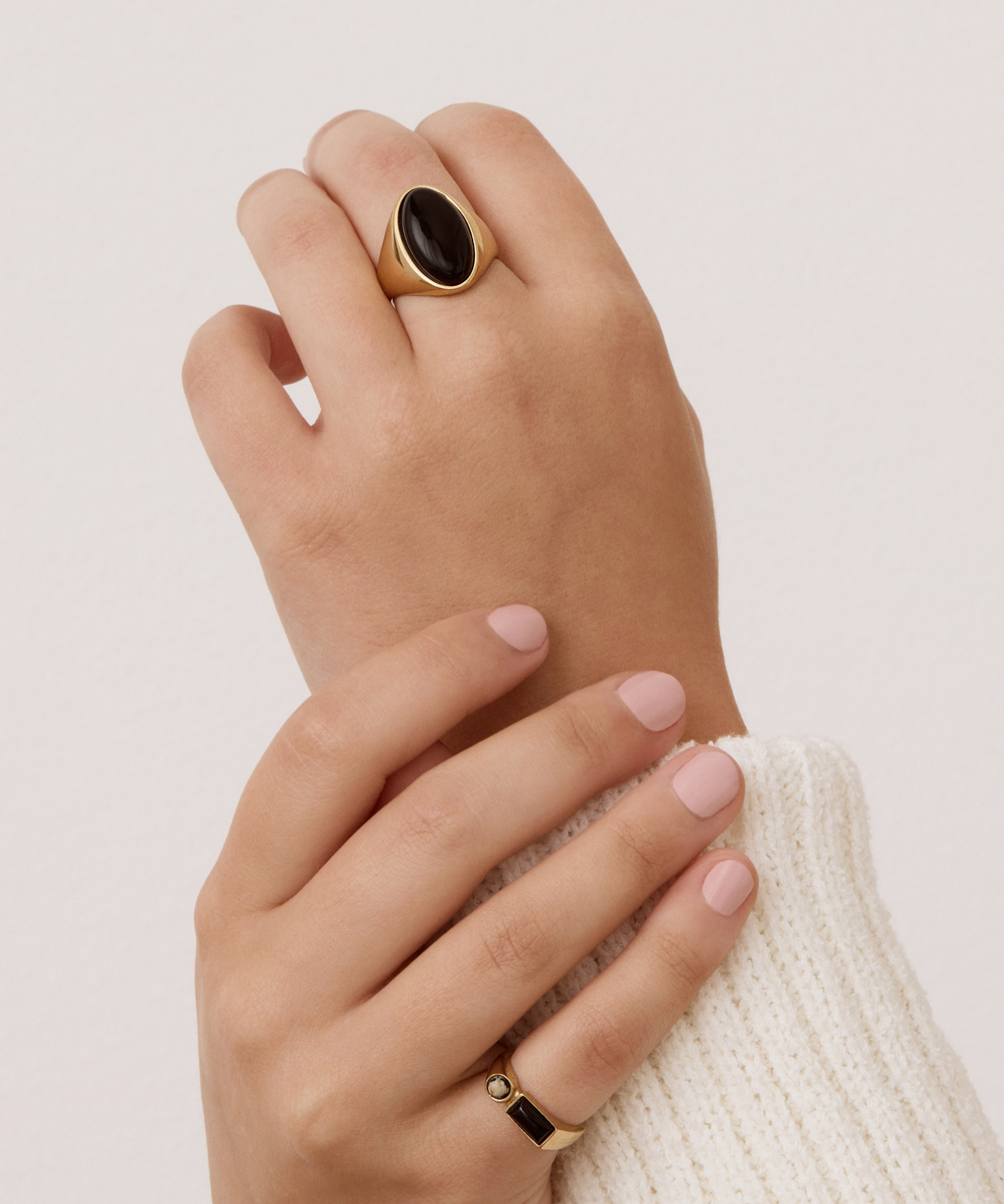 A close-up of a person's hands wearing gold rings, with the larger ring featuring a dark oval stone on the back hand and a smaller gold band with a black stone on the front hand, set against a plain light background.