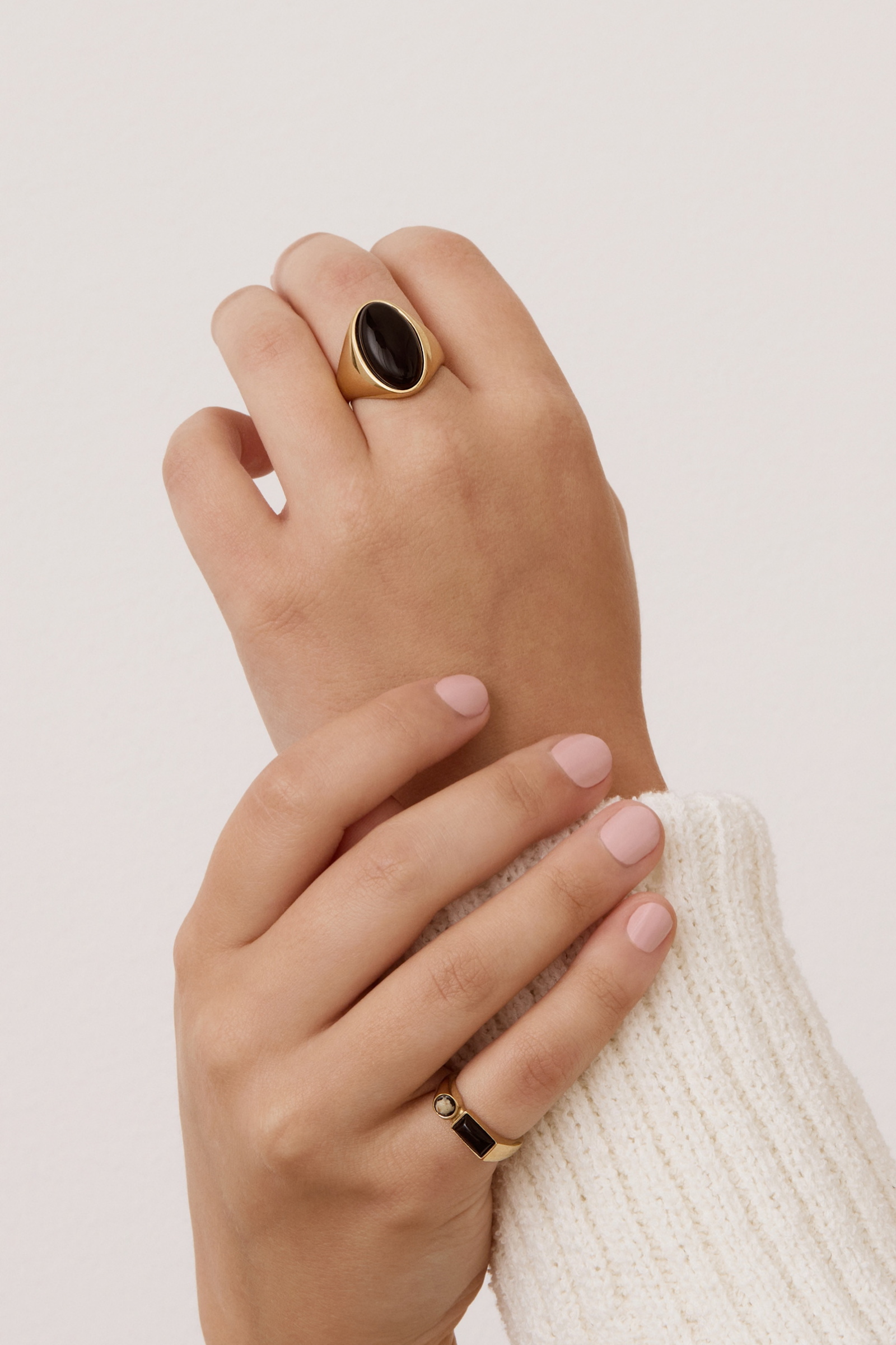 A close-up of a person's hands wearing gold rings, with the larger ring featuring a dark oval stone on the back hand and a smaller gold band with a black stone on the front hand, set against a plain light background.