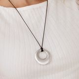 A close-up of a person wearing a silver circular pendant necklace on a black cord, paired with a white ribbed top.