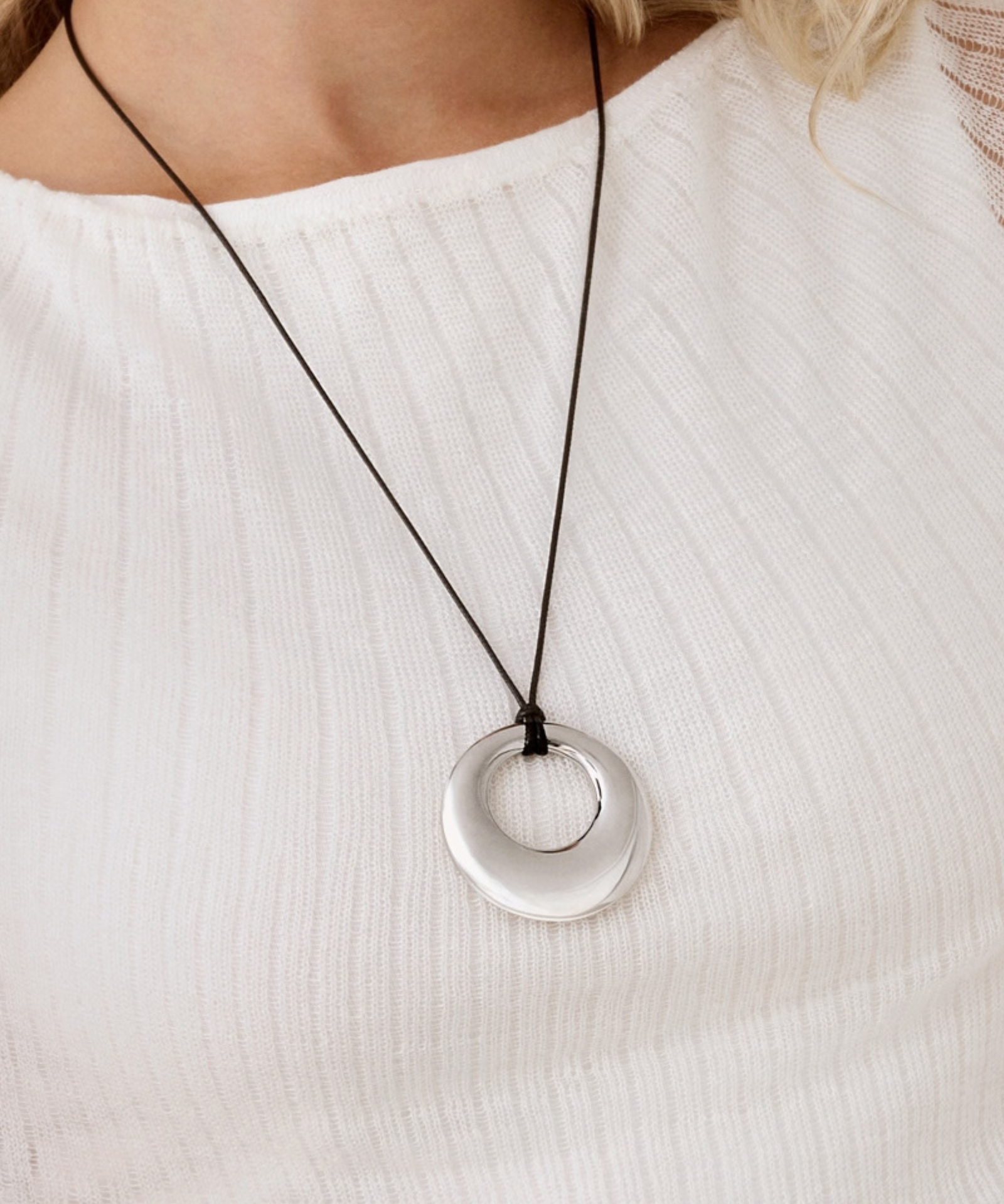 A close-up of a person wearing a silver circular pendant necklace on a black cord, paired with a white ribbed top.