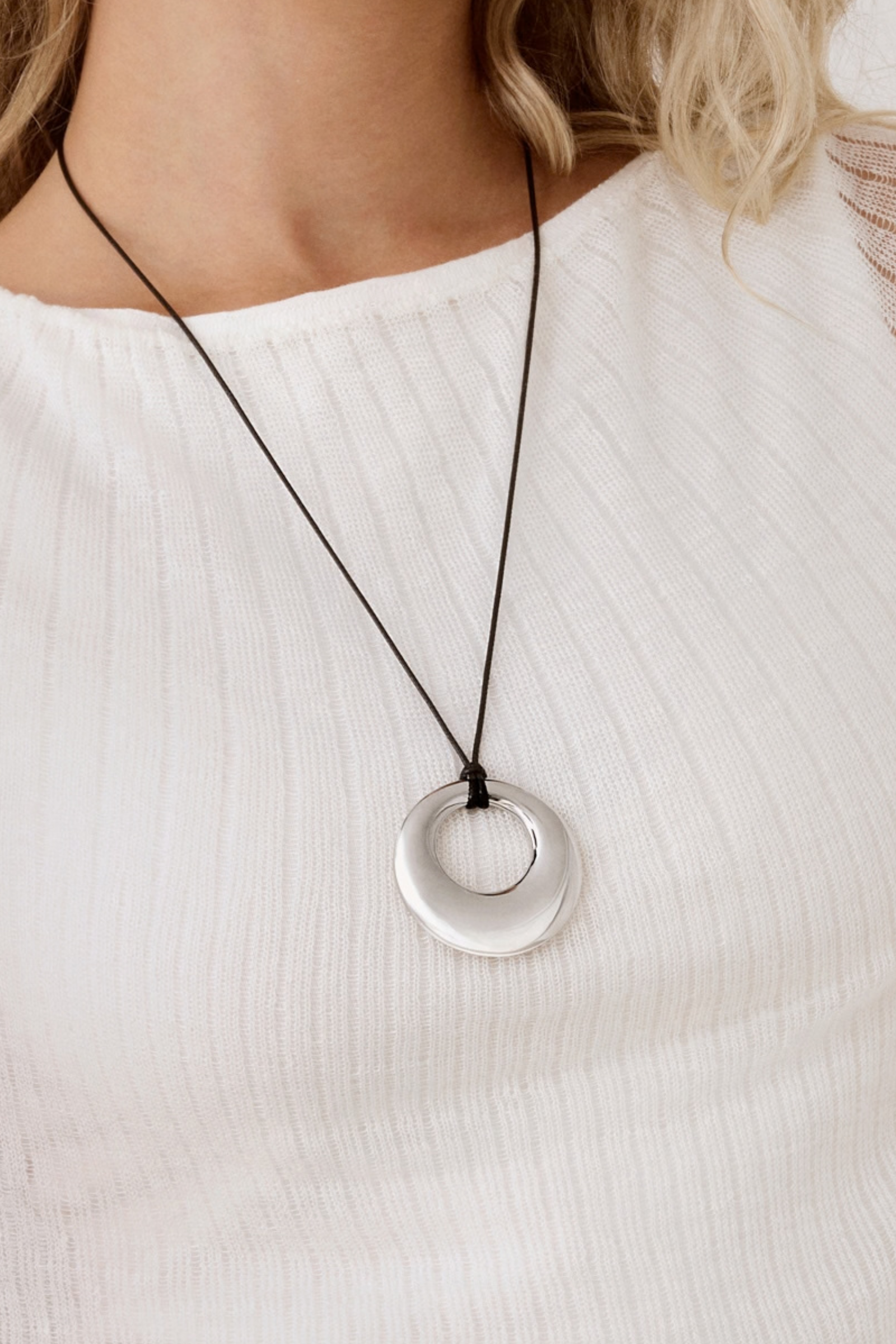 A close-up of a person wearing a silver circular pendant necklace on a black cord, paired with a white ribbed top.