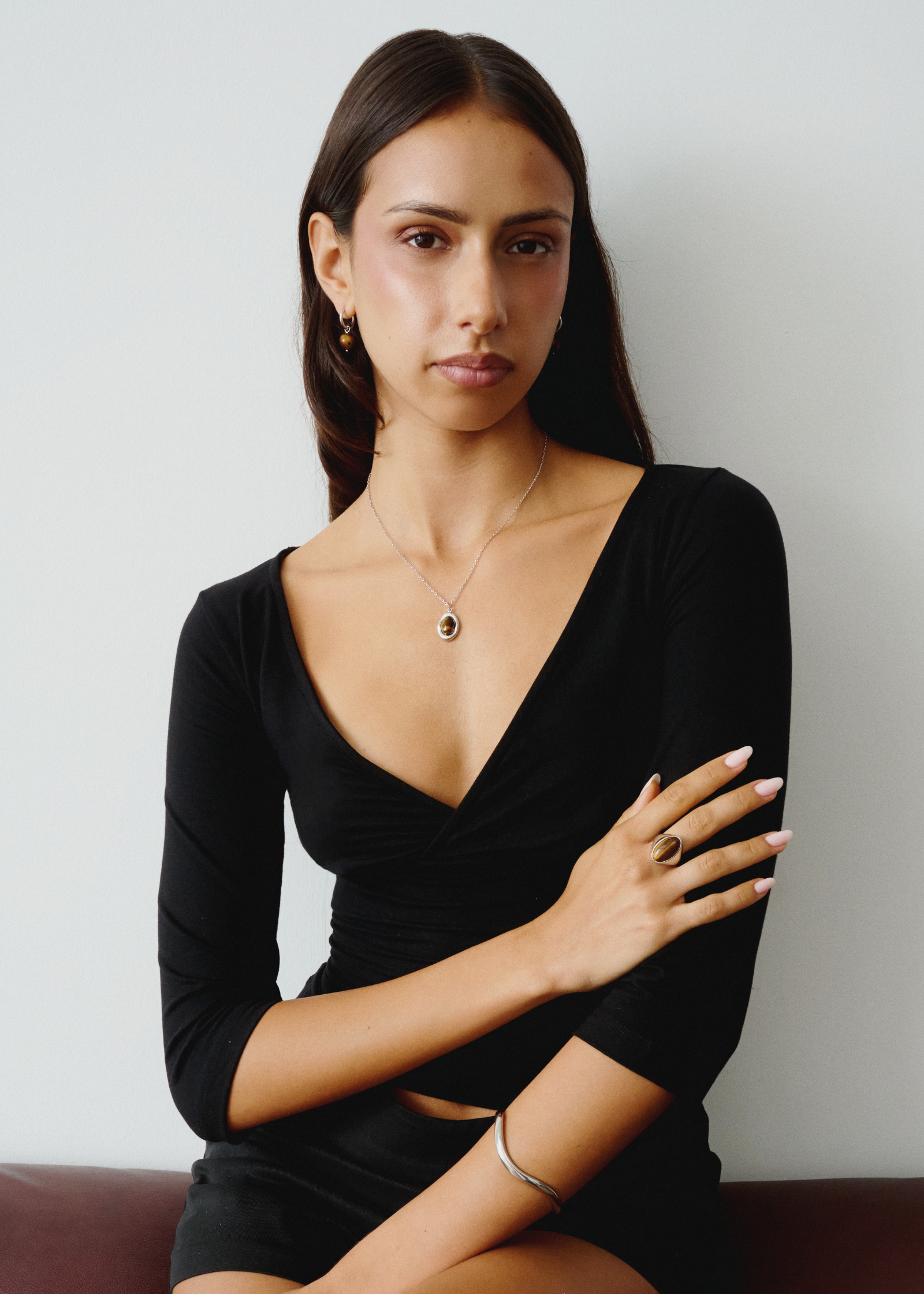 A woman with dark hair sits against a light gray wall wearing a black long-sleeve V-neck top and black shorts, accessorized with a delicate gold necklace and ring.