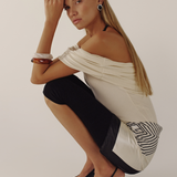 Woman in a white off-shoulder top and black pants posing against a plain background