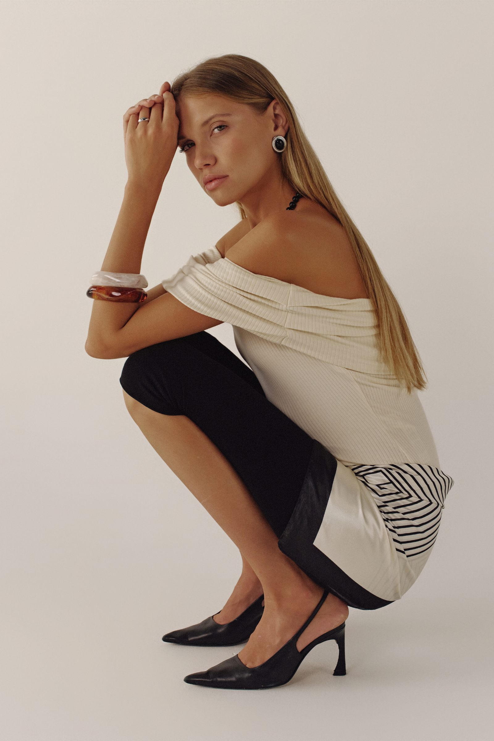 Woman in a white off-shoulder top and black pants posing against a plain background
