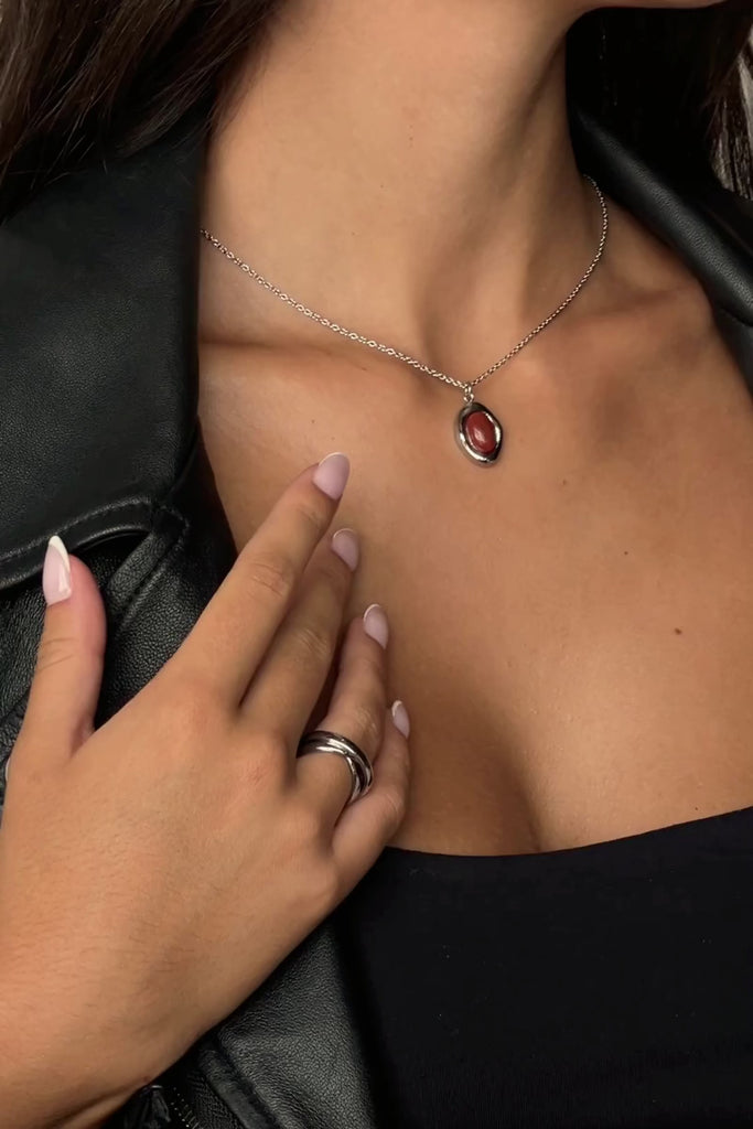 Video showcase of the Silver Bordeaux Necklace by Twenty Compass. The video features close-up shots of the organic-shaped Red Jasper stone pendant and the high-shine silver stainless steel chain. It demonstrates the adjustable 16 to 18-inch length and highlights the unique, natural variations in the deep red gemstone.