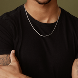 A person wearing a Twenty Compass 2mm Rope Necklace in silver and a black t-shirt crosses their tattooed arms, enhancing their edgy style. The muted olive green background adds texture to the scene as their face remains partially hidden.