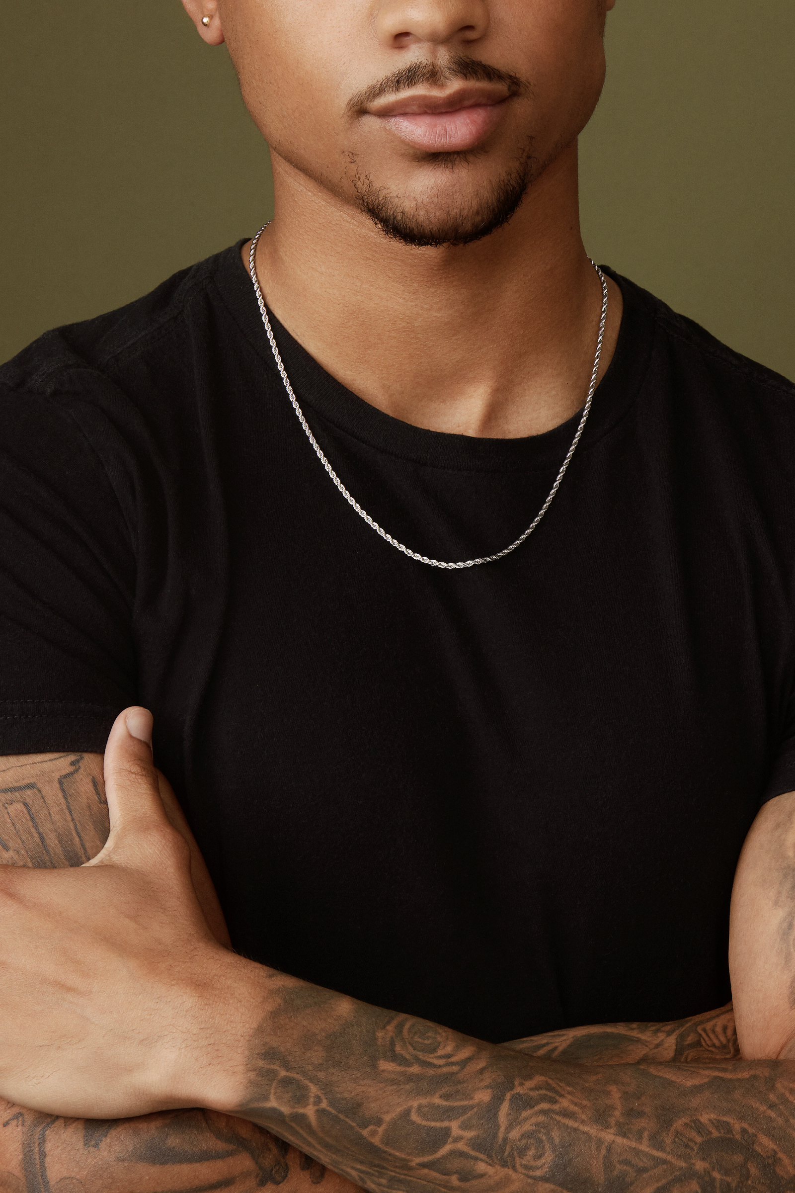 A person wearing a Twenty Compass 2mm Rope Necklace in silver and a black t-shirt crosses their tattooed arms, enhancing their edgy style. The muted olive green background adds texture to the scene as their face remains partially hidden.