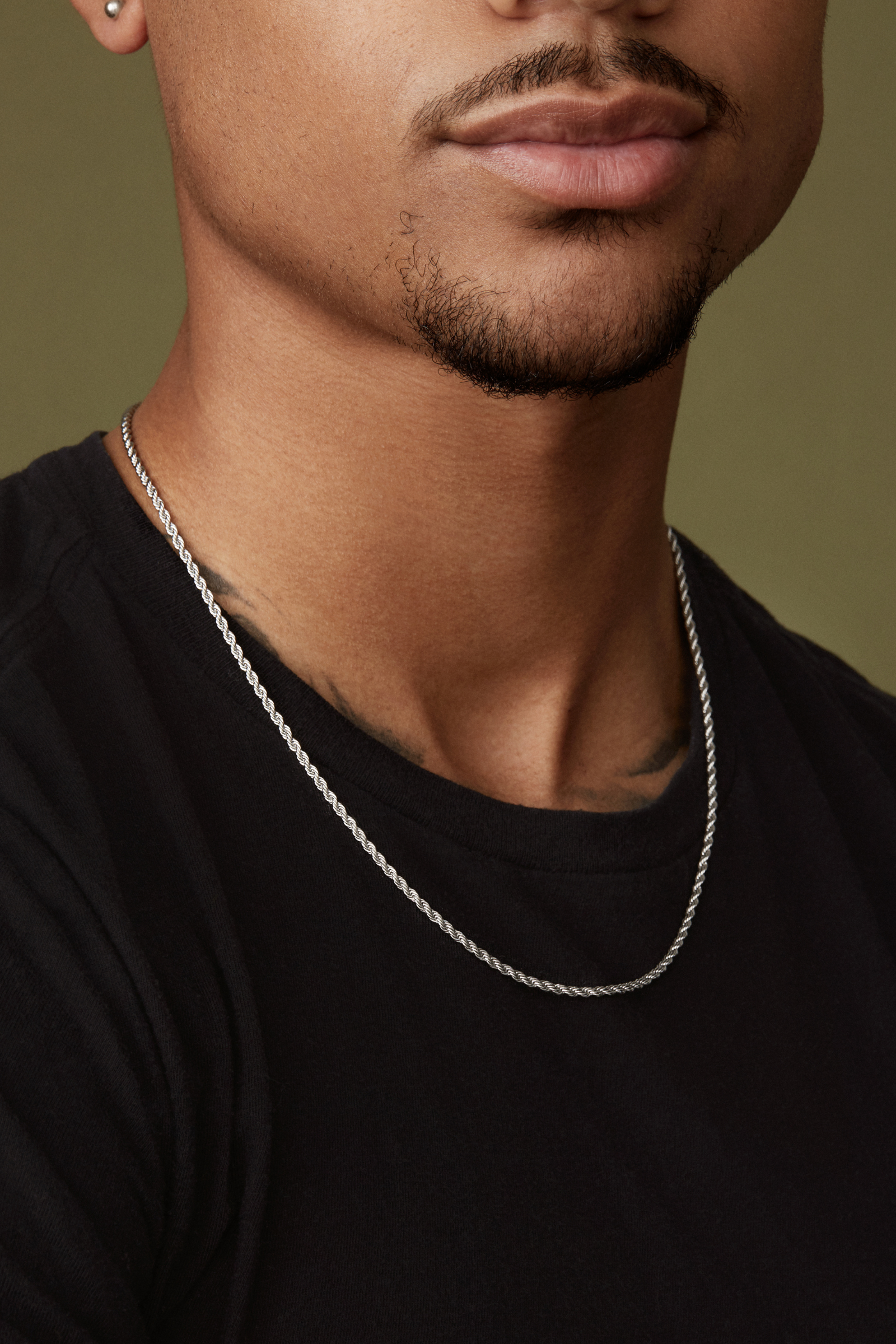A person wearing a black shirt and a Twenty Compass 2mm Rope Necklace in silver stands against an olive-green backdrop. The composition reveals only the lower face and neck, adding intrigue while highlighting the accessorys subtle style and texture.