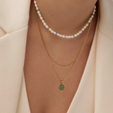 The image highlights someone wearing a pearl choker with colorful beads and the Andy Necklace by Twenty Compass, featuring a gold chain with a round green aventurine stone pendant. They are elegantly dressed in a cream-colored blazer.