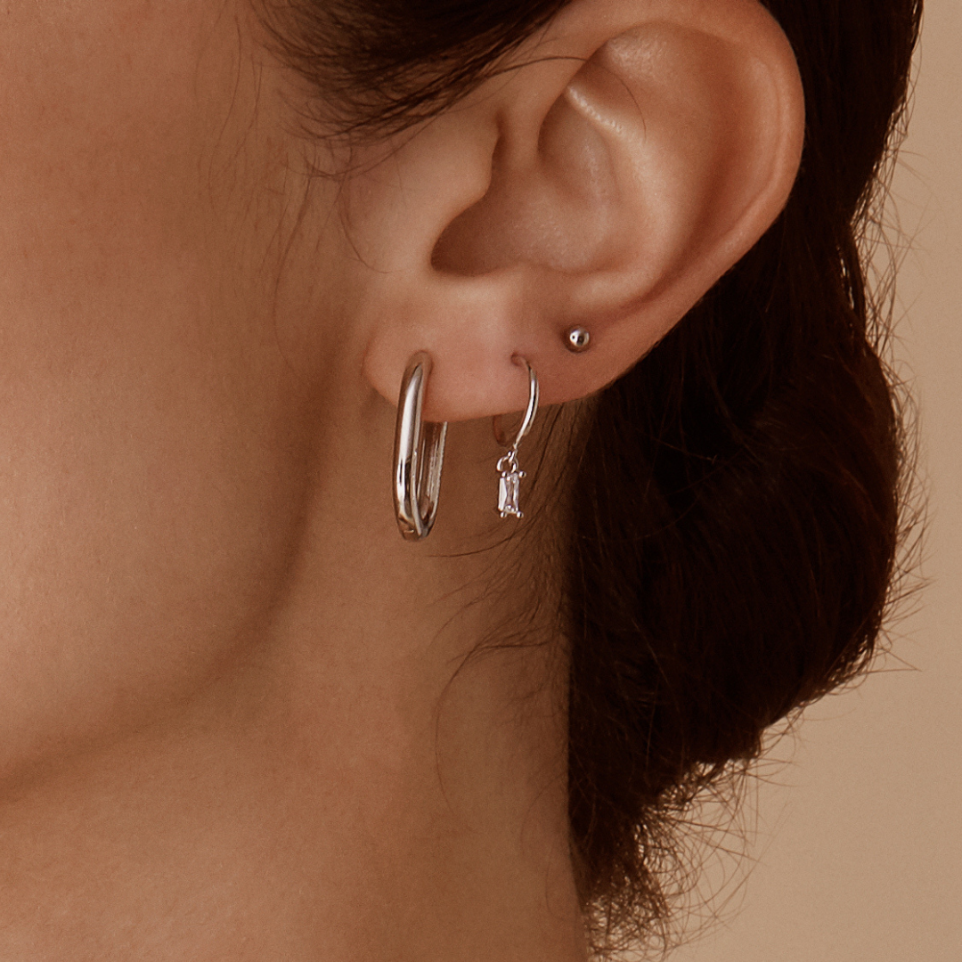 Close-up of a persons ear wearing two hoop earrings; the larger one, Alaska Earrings - Silver by Twenty Compass, is minimalist silver. The smaller hoop sparkles with a dangling cubic zirconia jewel against a soft beige background.