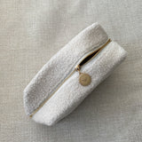 A white travel pouch with a textured surface, closed with a zipper featuring a gold-tone pendant with an engraved butterfly design.