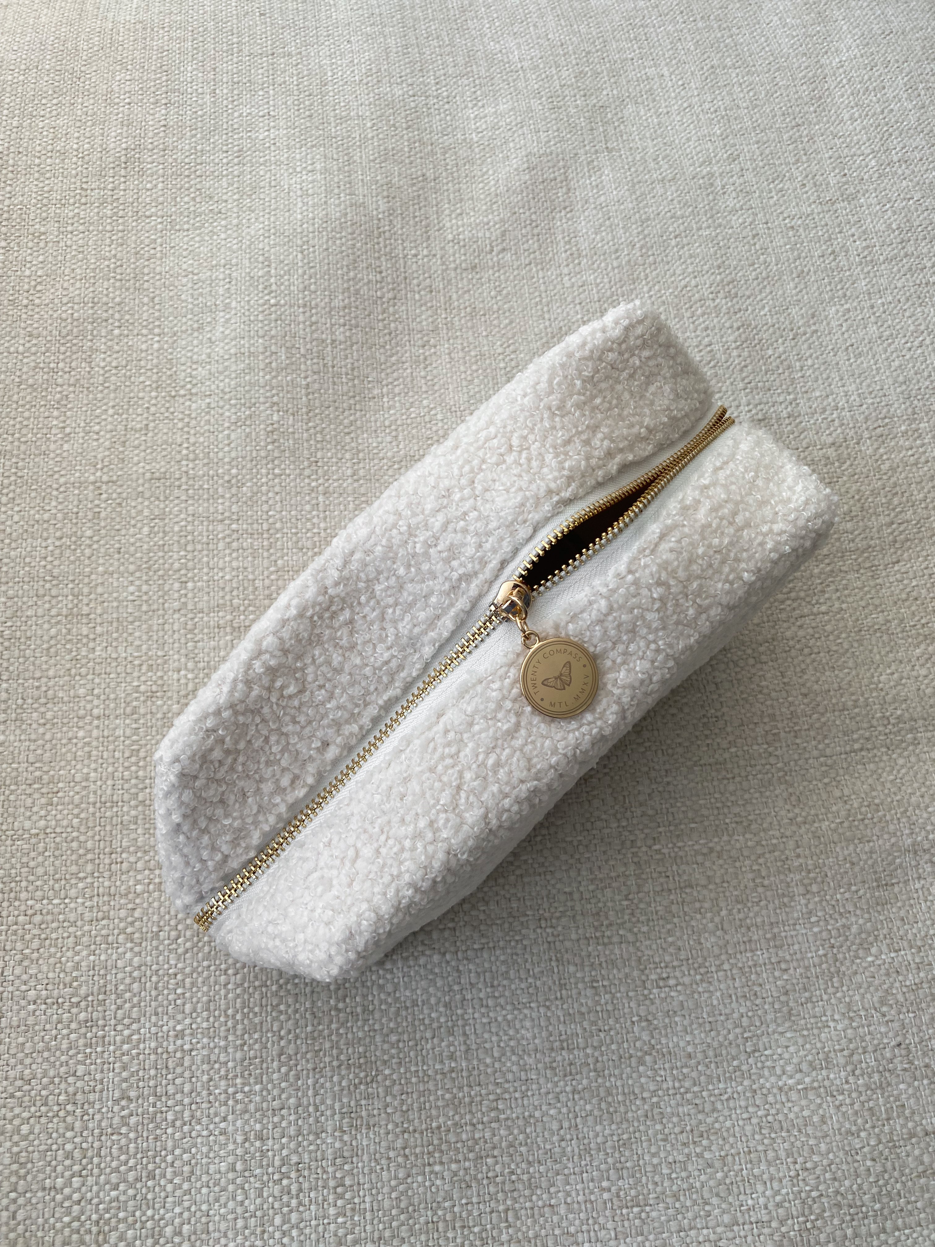 A white travel pouch with a textured surface, closed with a zipper featuring a gold-tone pendant with an engraved butterfly design.