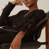 A person with short hair sits relaxed on a black chair, wearing a minimalist black outfit and Alaska Earrings - Silver by Twenty Compass. Gold jewelry glints as one hand rests on their face and the other on the armrest, while the neutral background highlights their confident expression.