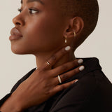 A person with a shaved head and wearing the Alaska Ring in Gold Vermeil by Twenty Compass glances to the side, hand touching their neck. Theyre in a black outfit, showcasing neatly manicured white nails that glisten like cubic zirconia.